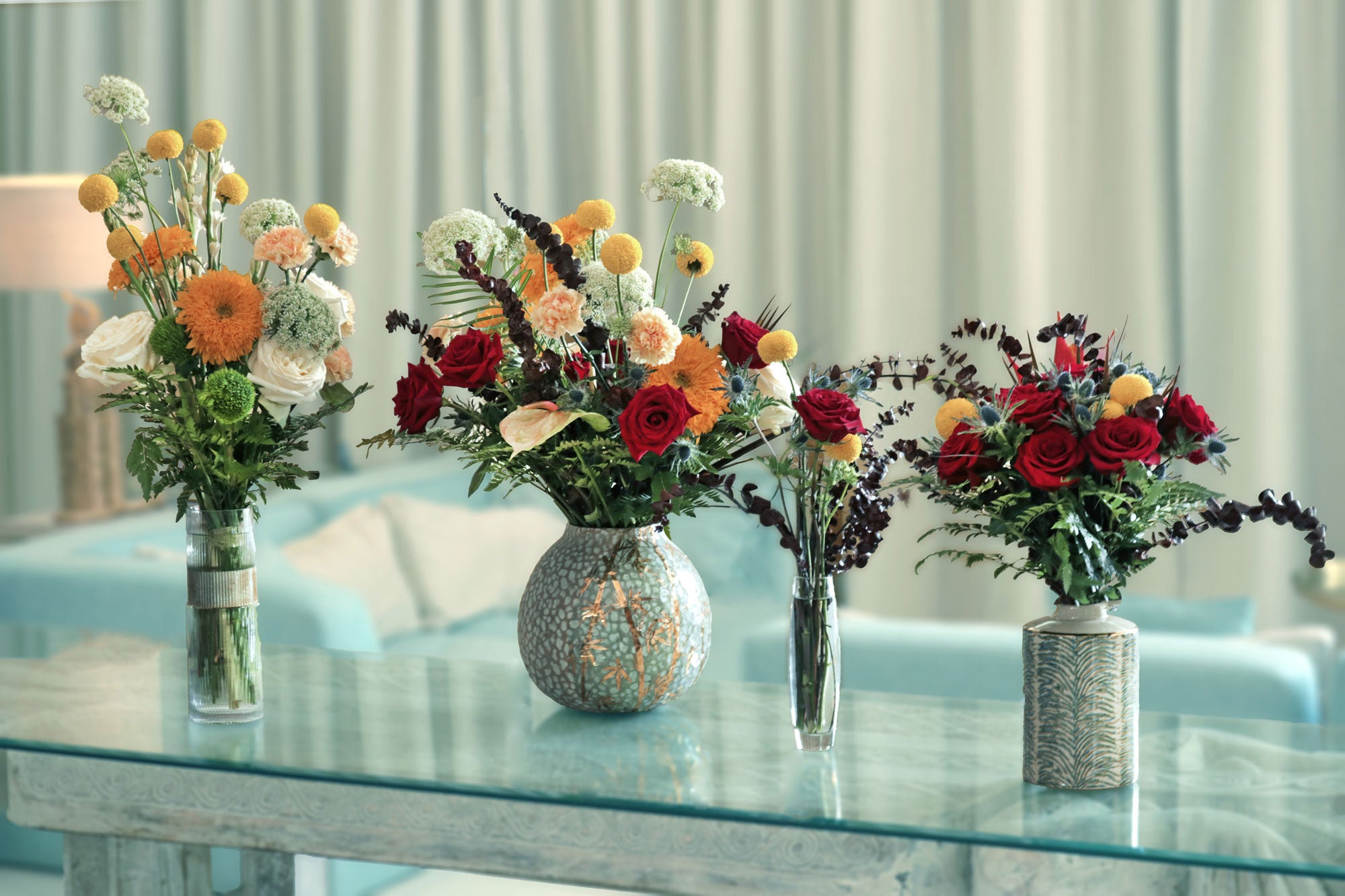 4 different sized vases with fresh flowers arranged in decorative bouquets on a glass table.
