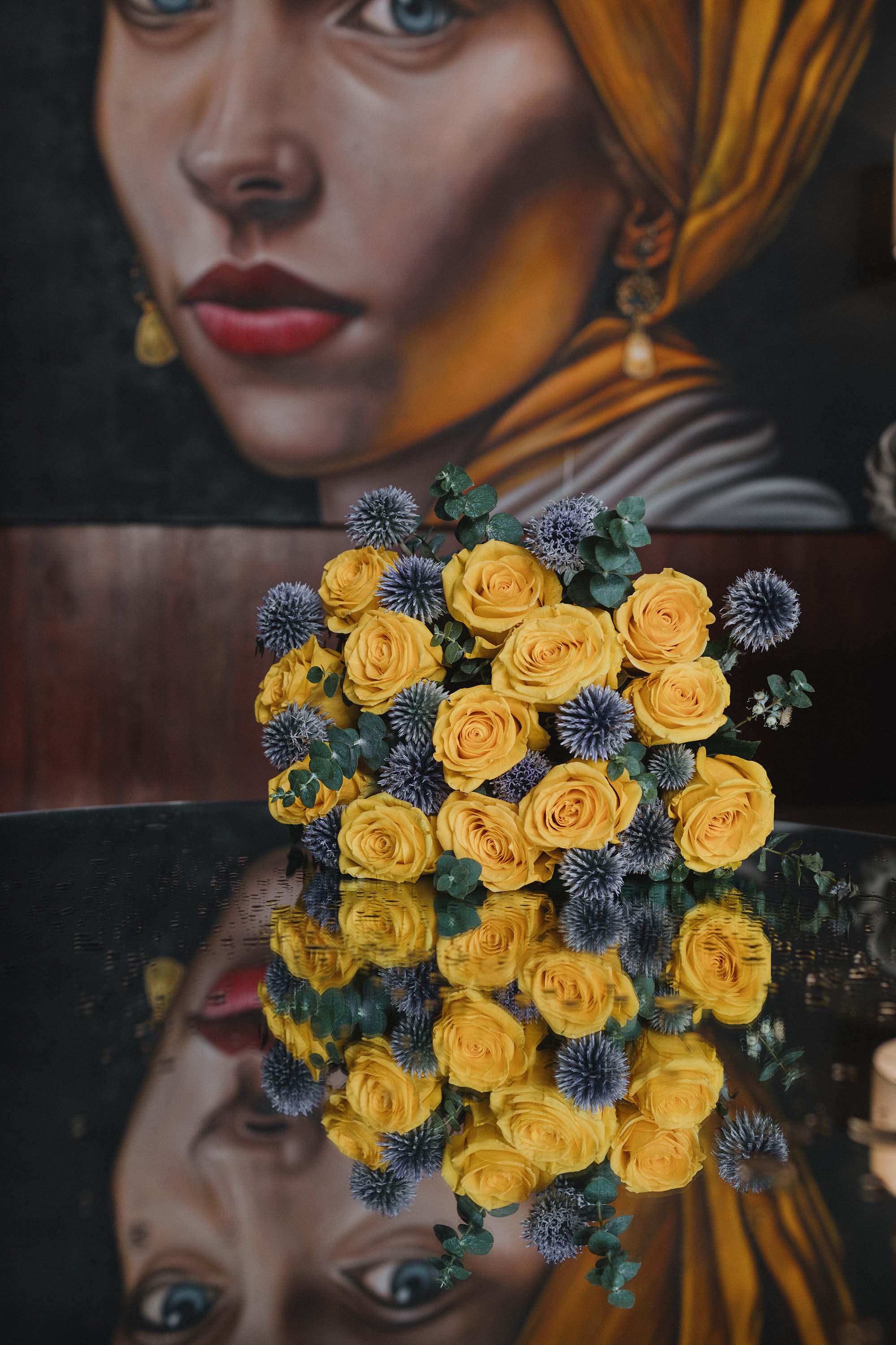 Bouquet of yellow roses and blue flowers in front of a painting of a woman with a yellow headscarf.
