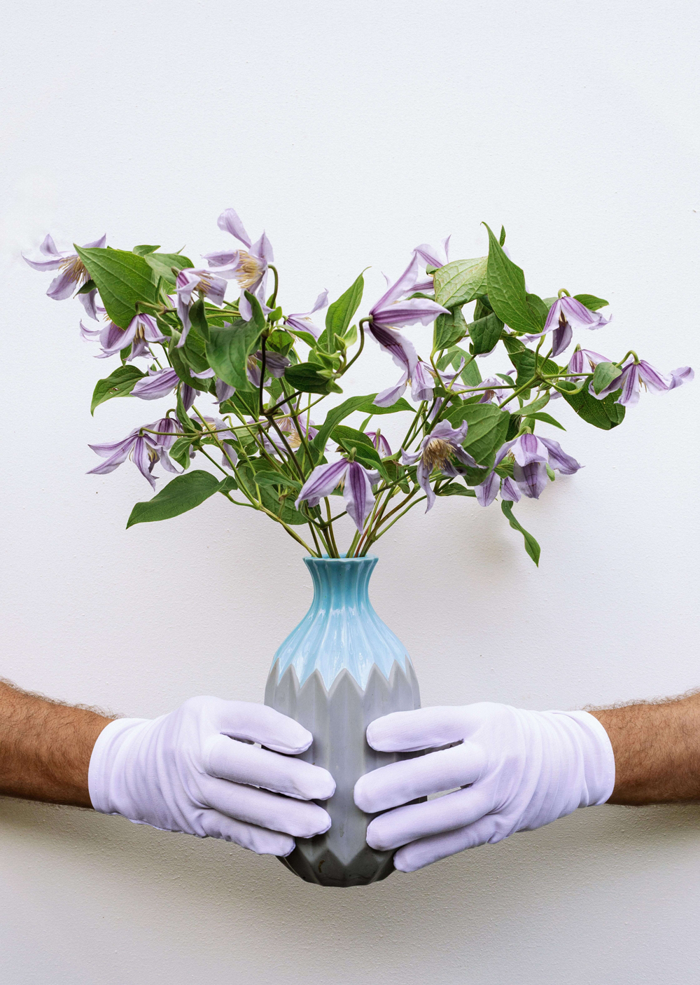Person holding a vase with purple flowers wearing white gloves on a light background